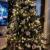 Tamnozeleno gusto umjetno božićno drvce, ukrašeno bijelo-ružičastim ukrasima i toplom bijelom rasvjetom, s bijelim tepihom ispod drvca