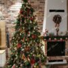 Visoko umjetno božićno drvce sa zelenim granama s ugrađenom LED rasvjetom, gusto ukrašeno crvenim i zelenim ukrasima, uz kamin u dnevnom boravku